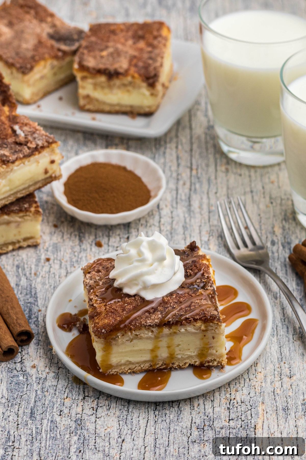 Churro Cheesecake Recipe: Cinnamon-Sugar Cheesecake Bars 2 Slice of cheesecake on a small white plate, drizzled with caramel syrup and topped with whipped cream.