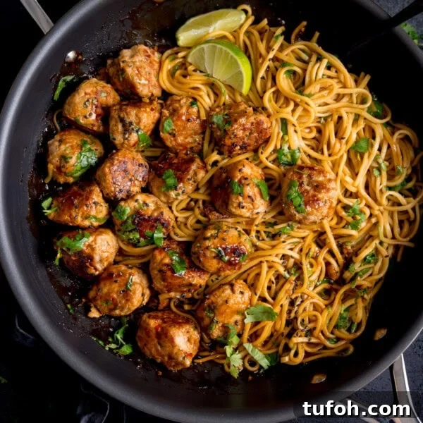 Cooked noodles and browned lemongrass meatballs in a frying pan with herbs and lime wedges.