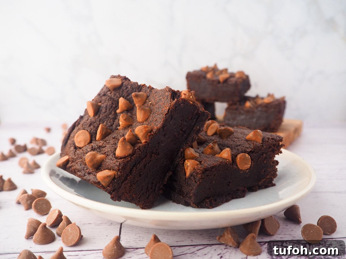 Decadent Egg-Free Brownies: Fudgy, Moist Recipe 15 Two eggless brownies on a plate, with a stack of eggless brownies in the background, and chocolate chips scattered around.