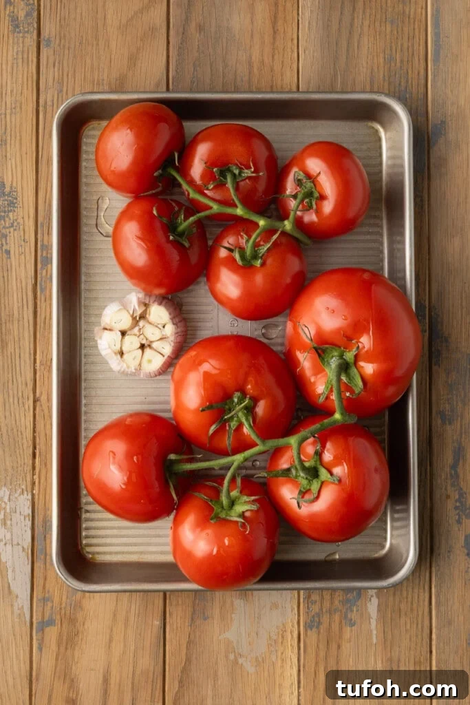 A sheet pan with a head of garlic with the top cut off and ten tomatoes on the vine, all covered in olive oil. 