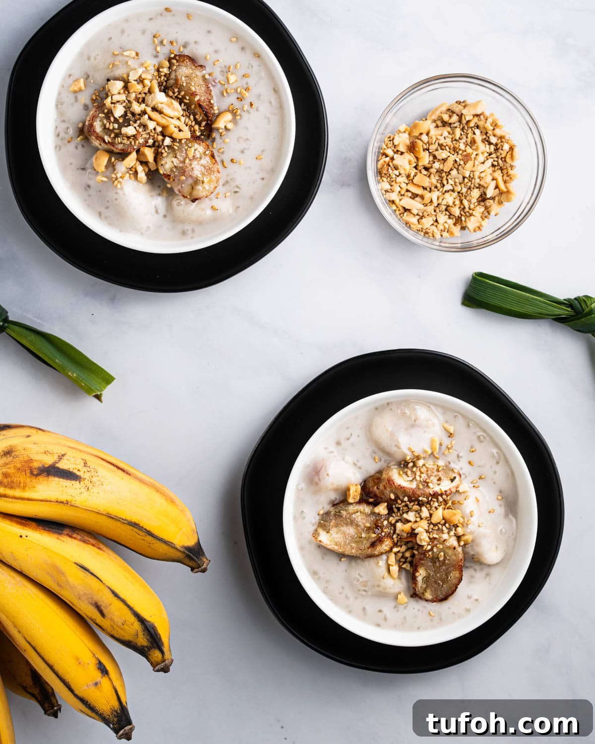 two bowls of che chuoi surrounded by bananas, pandan leaves tied in knots, and a bowl of crushed peanuts and sesame seeds.