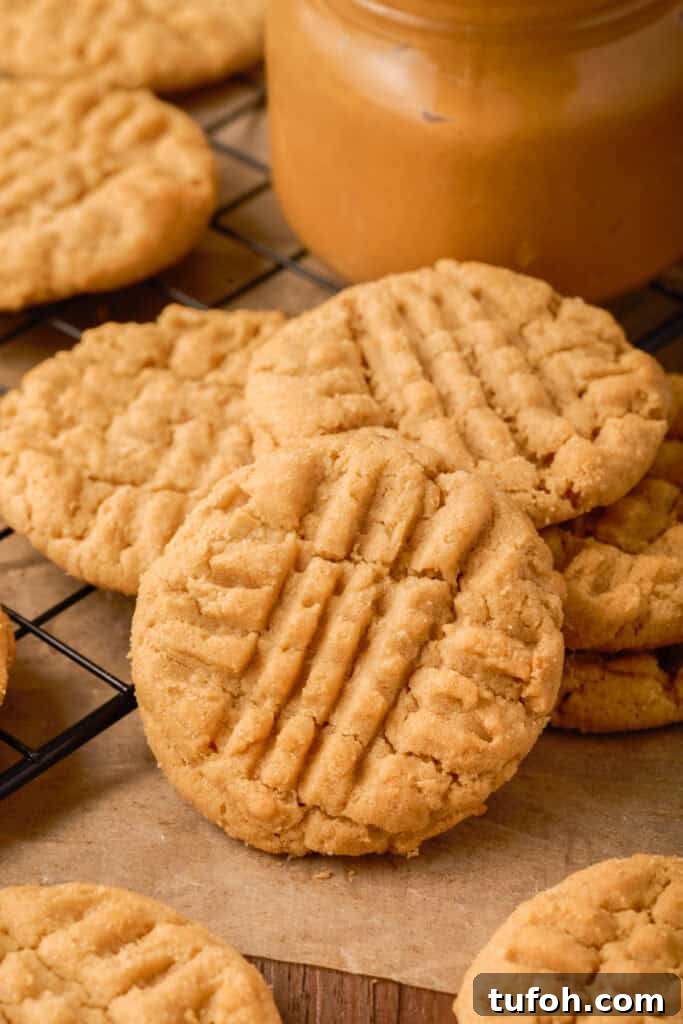 Chewy Peanut Butter Cookies Recipe with Crispy Edges 2 Stack of soft peanut butter cookies cooling near a jar of peanut butter and a red-striped kitchen towel.
