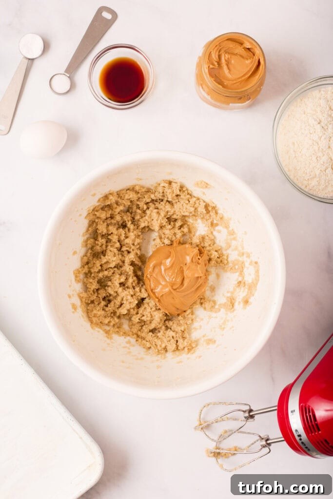 Chewy Peanut Butter Cookies Recipe with Crispy Edges 6 Peanut butter added to the creamed butter and sugar mixture in a white bowl with remaining ingredients and tools visible on the counter.