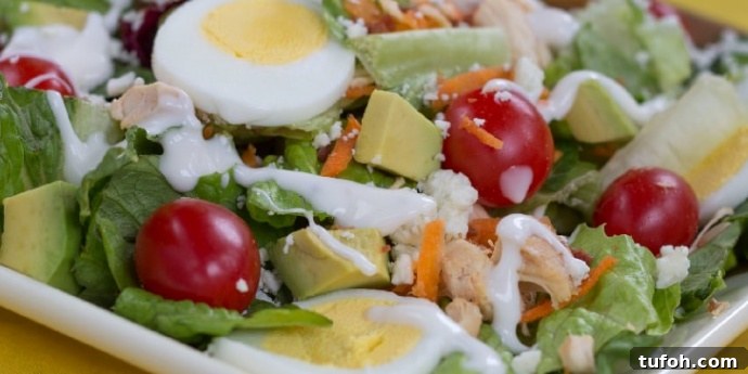Cobb Salad with Chicken and Avocado Cobb salad on a white square plate on a yellow tablecloth. Cobb salad ingredients are lettuce, grape tomatoes, sliced boiled eggs, diced avocado, diced chicken, shredded carrots, and blue cheese dressing.