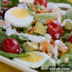Chicken and Avocado Cobb Salad Recipe for Lunch or Dinner 2 Cobb salad on a white square plate on a yellow tablecloth. Cobb salad ingredients are lettuce, grape tomatoes, sliced boiled eggs, diced avocado, diced chicken, shredded carrots, and blue cheese dressing.