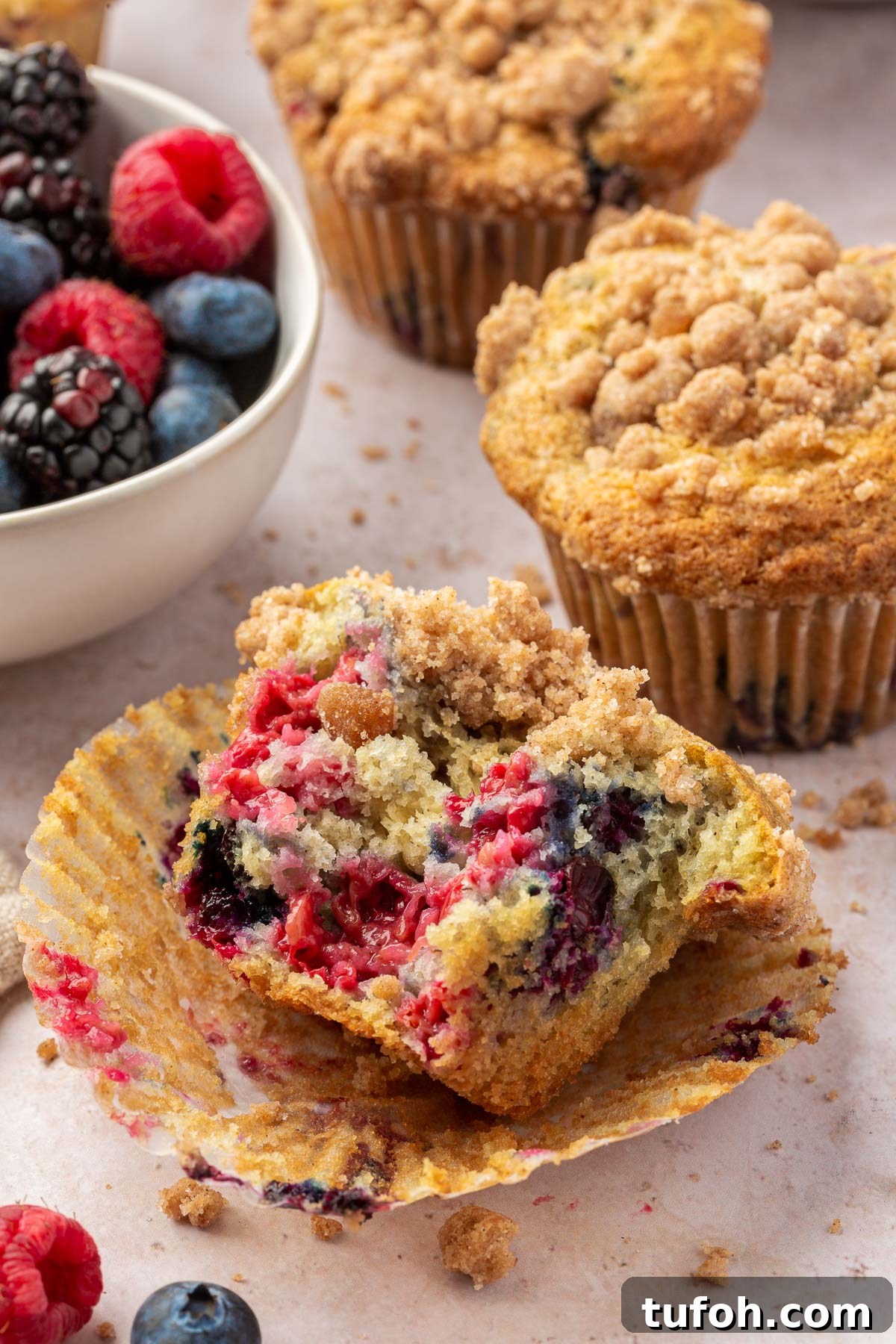 Gluten-Free Mixed Berry Muffins Recipe — Light, Moist & Berry-Packed 4 Half of a gf triple berry muffin that has been bitten into and is laying on the muffin liner with additional muffins and streusel crumbs in the background.