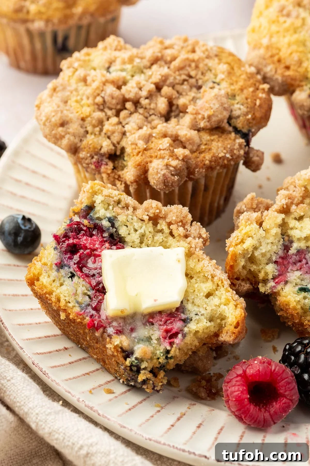 Gluten-Free Mixed Berry Muffins Recipe — Light, Moist & Berry-Packed 5 Half of a gluten free mixed berry muffin on a plate with a pat of melted butter on it with additional muffins and fresh berries on the plate.