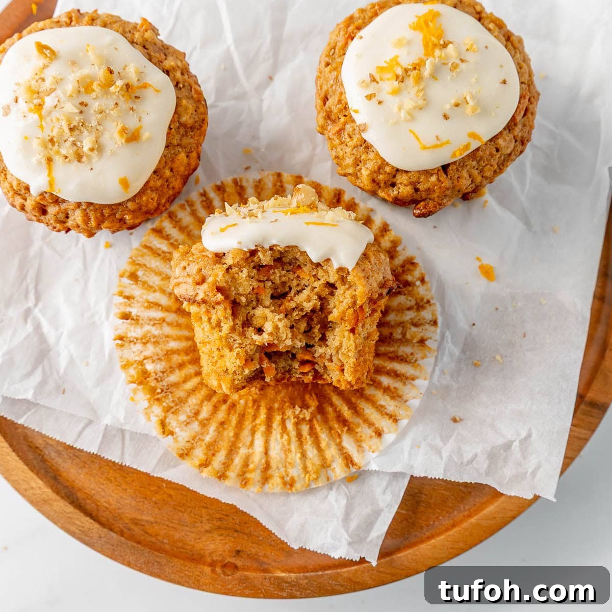 Moist Carrot Cake Muffins with Cream Cheese Swirl 12 Three carrot muffins with frosting sit on parchment paper on a wooden plate. One muffin has a bite taken out, revealing its moist interior.