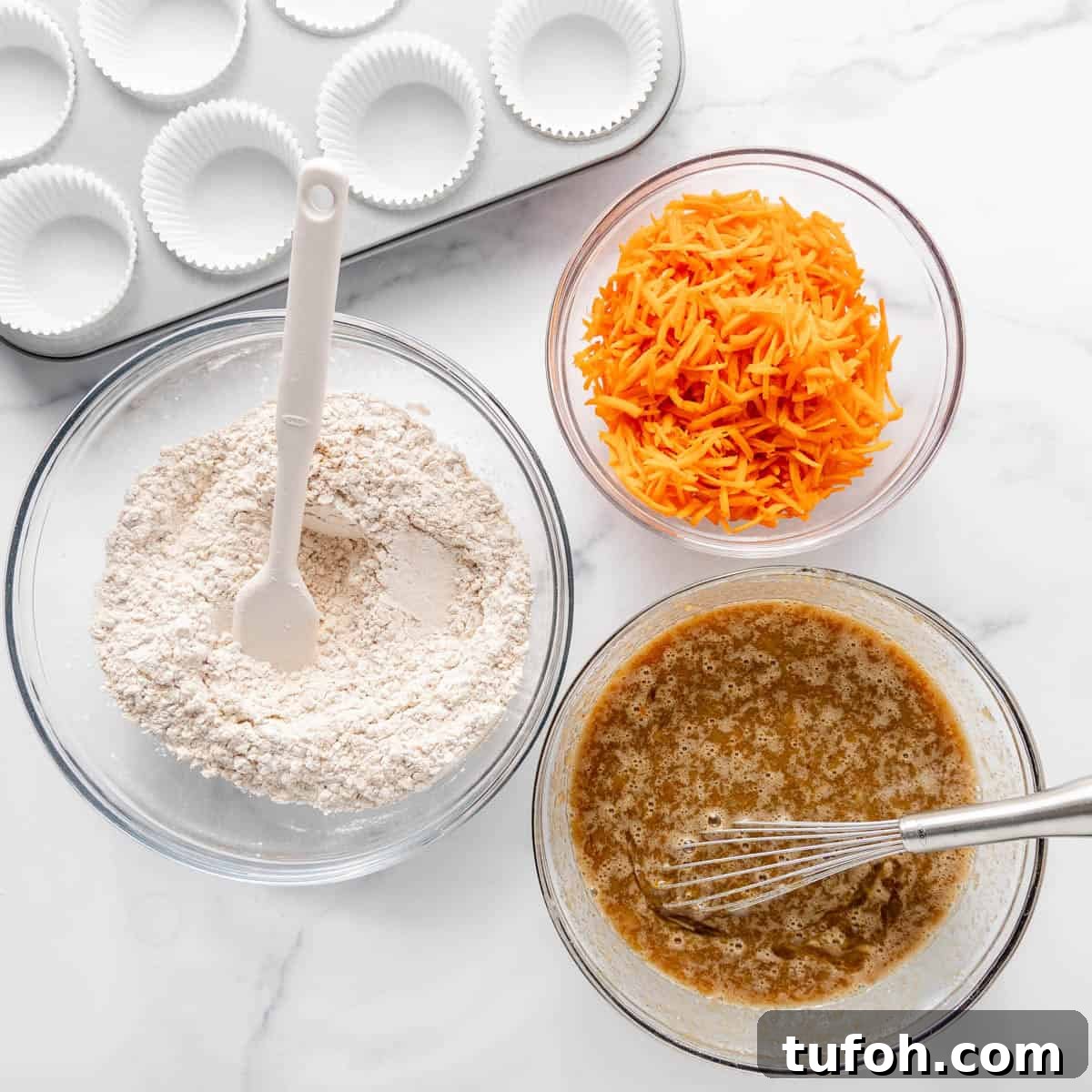 Moist Carrot Cake Muffins with Cream Cheese Swirl 4 Three bowls on a marble surface: flour mix with spatula, shredded carrots, and wet ingredients with whisk. Cupcake tray with liners is in the background.