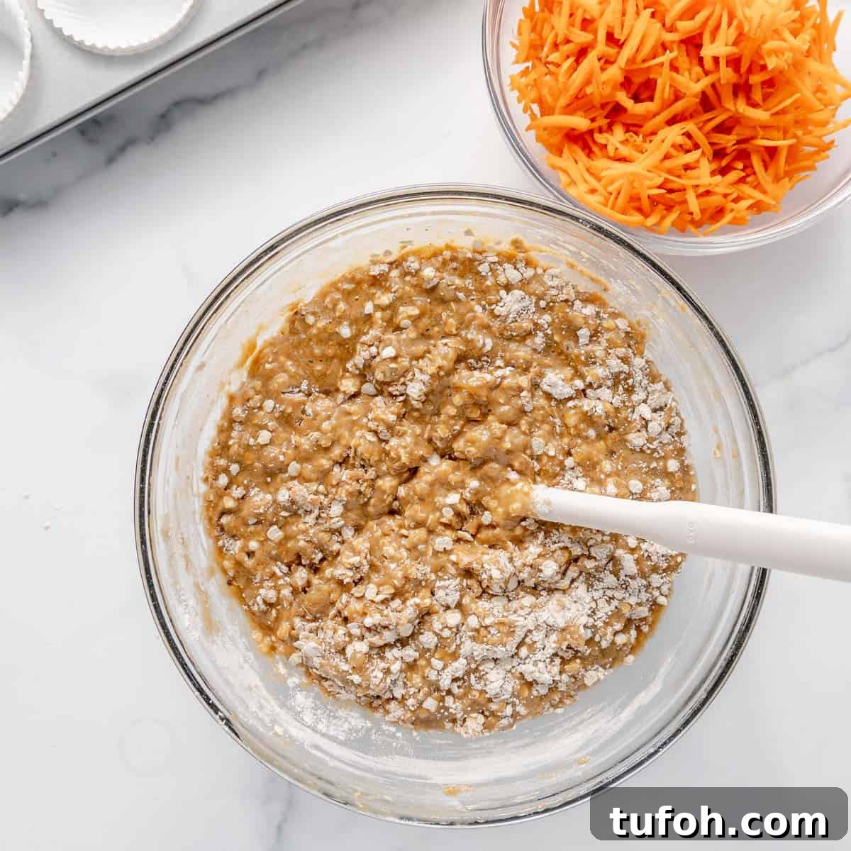 Moist Carrot Cake Muffins with Cream Cheese Swirl 5 Glass bowl with muffin batter being mixed, next to a bowl of shredded carrots on a white countertop.
