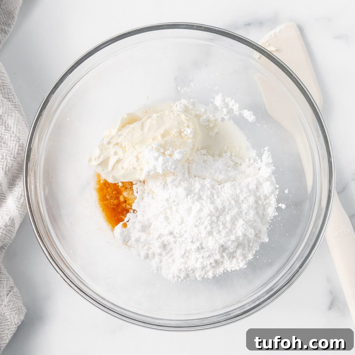 Moist Carrot Cake Muffins with Cream Cheese Swirl 8 A glass bowl contains cream cheese, powdered sugar, vanilla extract, and milk. A spatula is beside the bowl on a marble countertop.