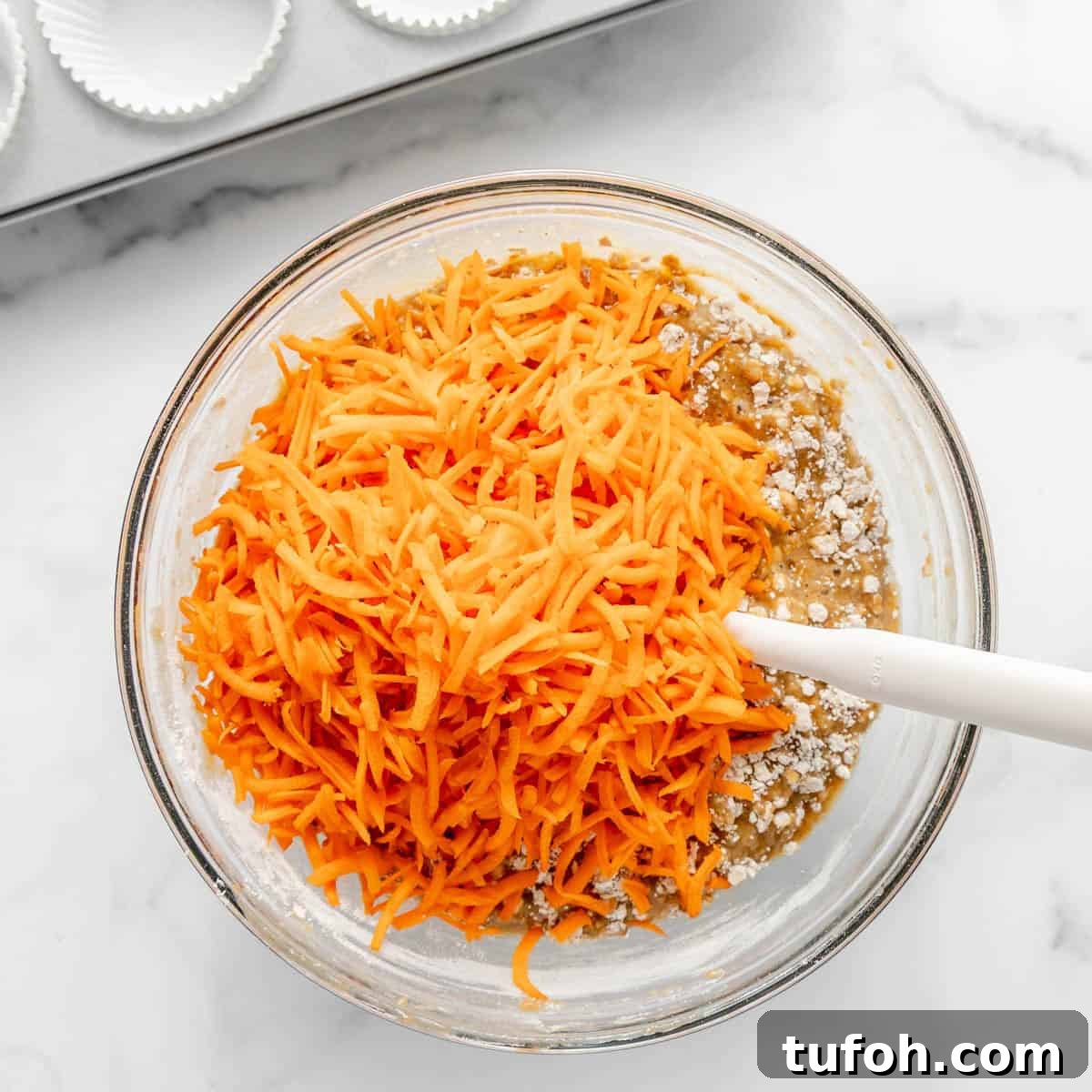 Moist Carrot Cake Muffins with Cream Cheese Swirl 10 A glass bowl with shredded carrots on top of dry ingredients and batter, being mixed with a white spatula. Batter appears to be for baking, likely muffins or cake.