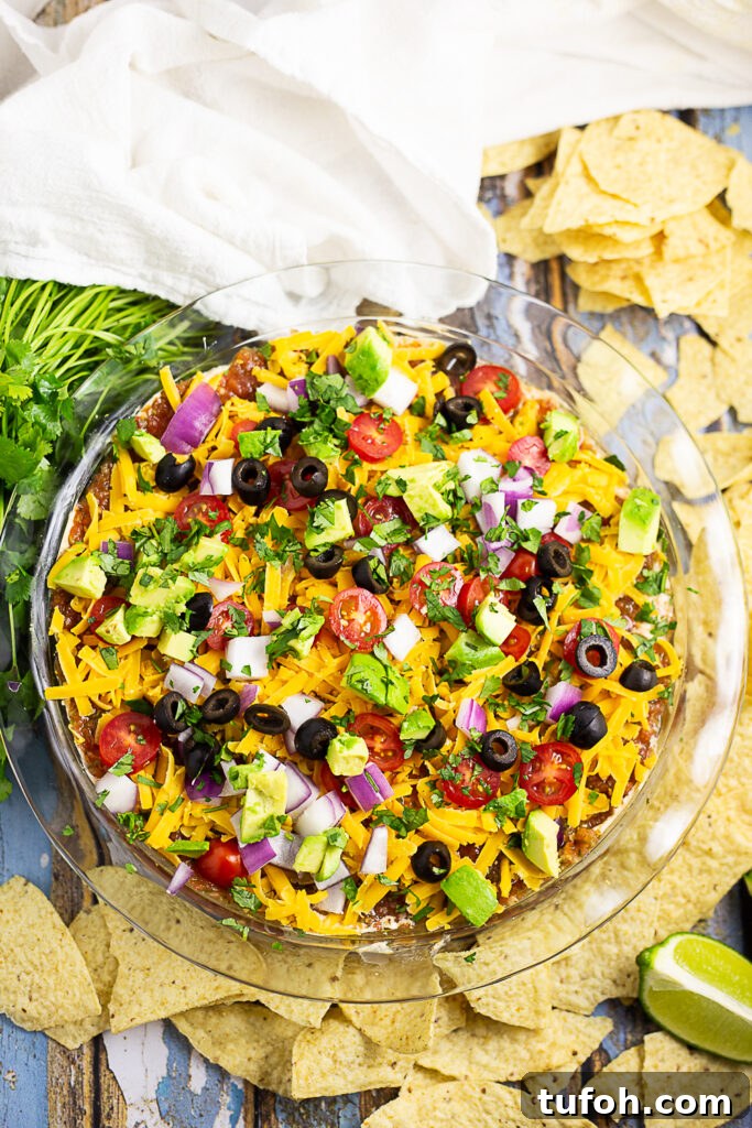 Overhead view of easy layered taco dip in a round glass dish surrounded by tortilla chips, a fresh bunch of cilantro, lime wedges and a clean linen.