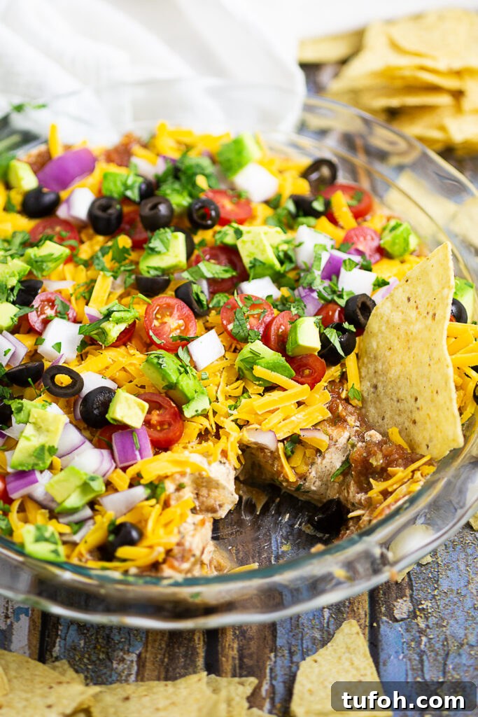 Layered taco dip in a round glass dish with a scoop missing and a chip sticking out of it. Tortilla chips and clean linen surround the dish