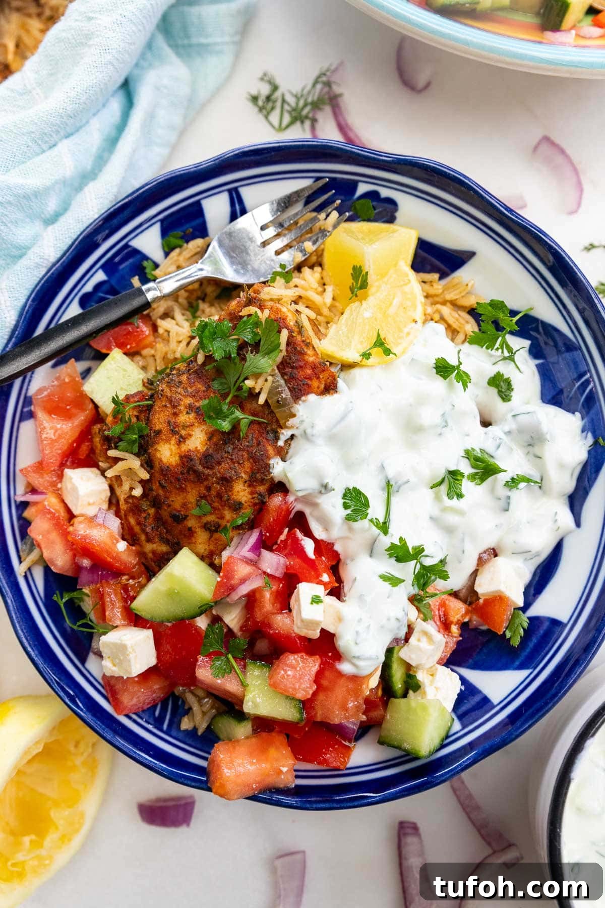 Closeup of overhead of a blue bowl of Greek chicken and rice with tomato, cucumber and feta salad and tsatziki.