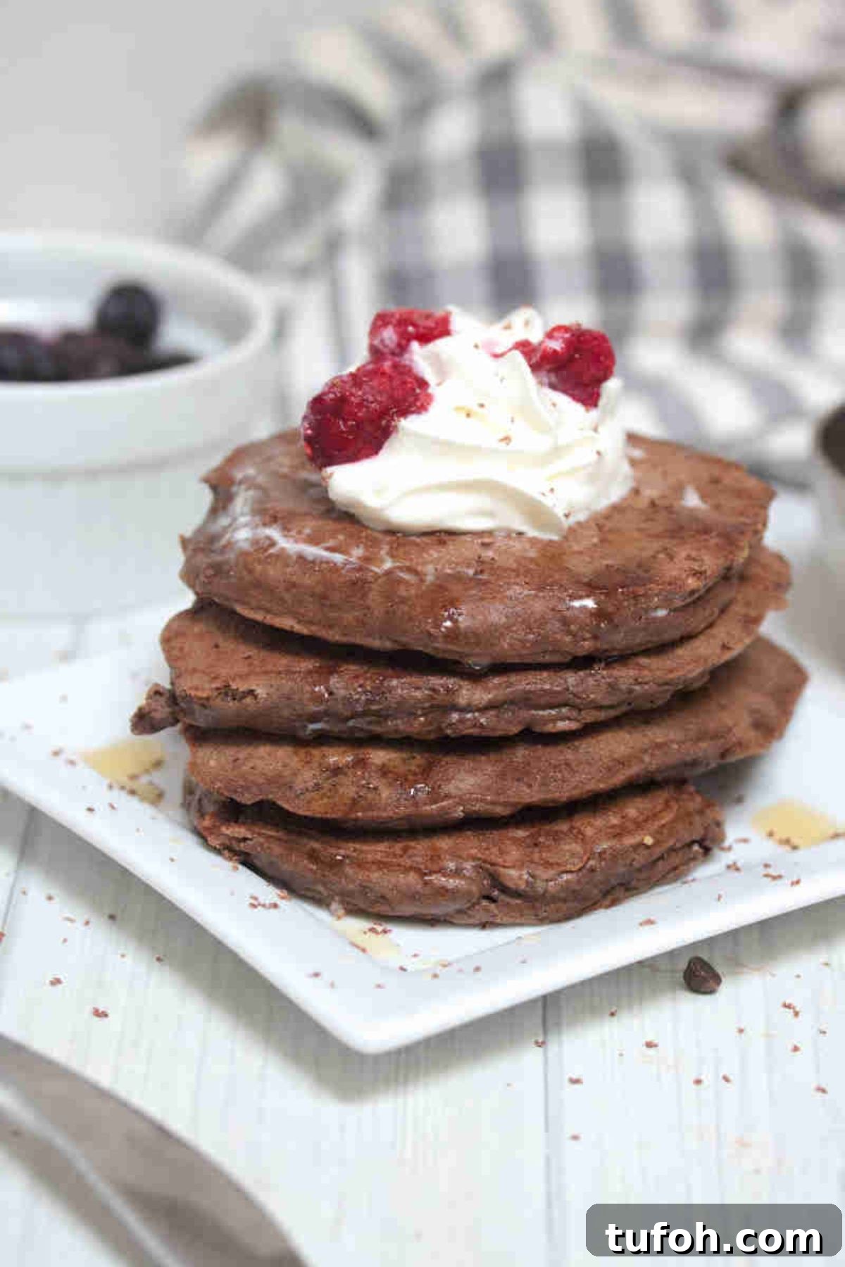 Keto chocolate pancakes stacked on a white plate and whipped cream and raspberries on top.