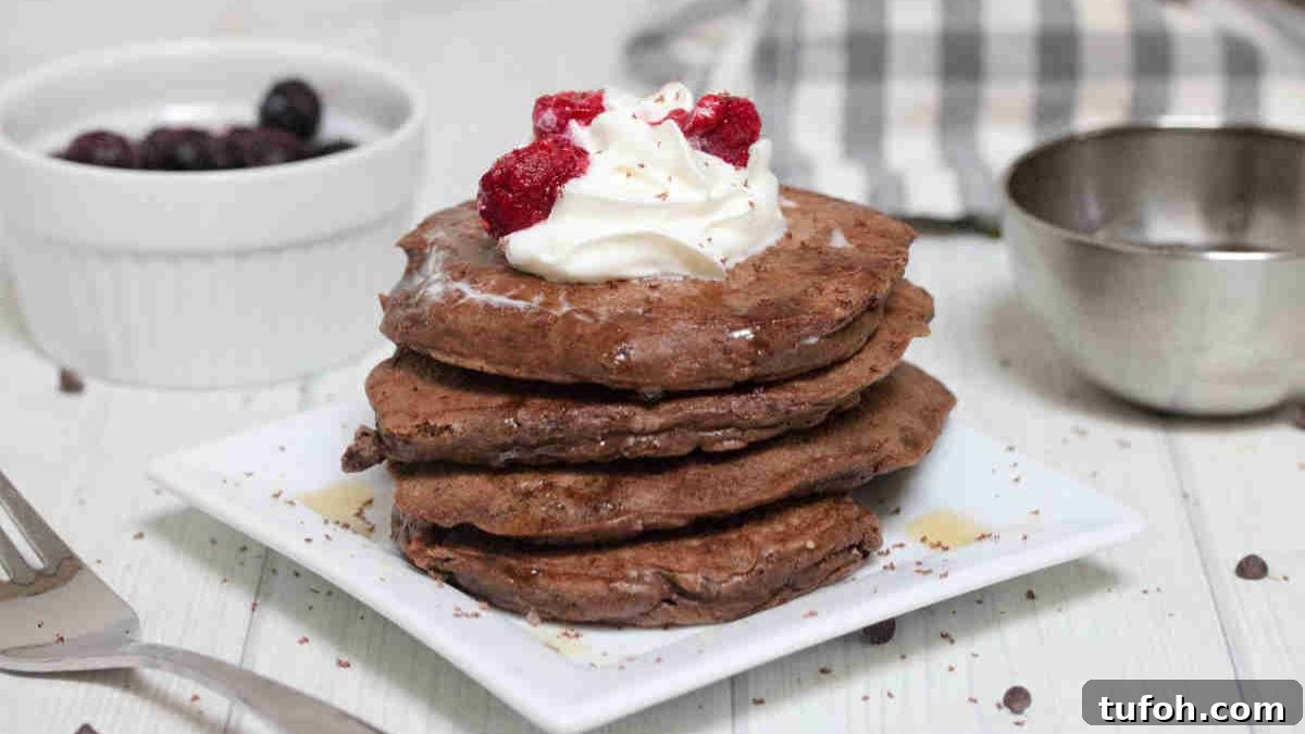 Keto chocolate pancakes stacked on a white plate and whipped cream and raspberries on top.