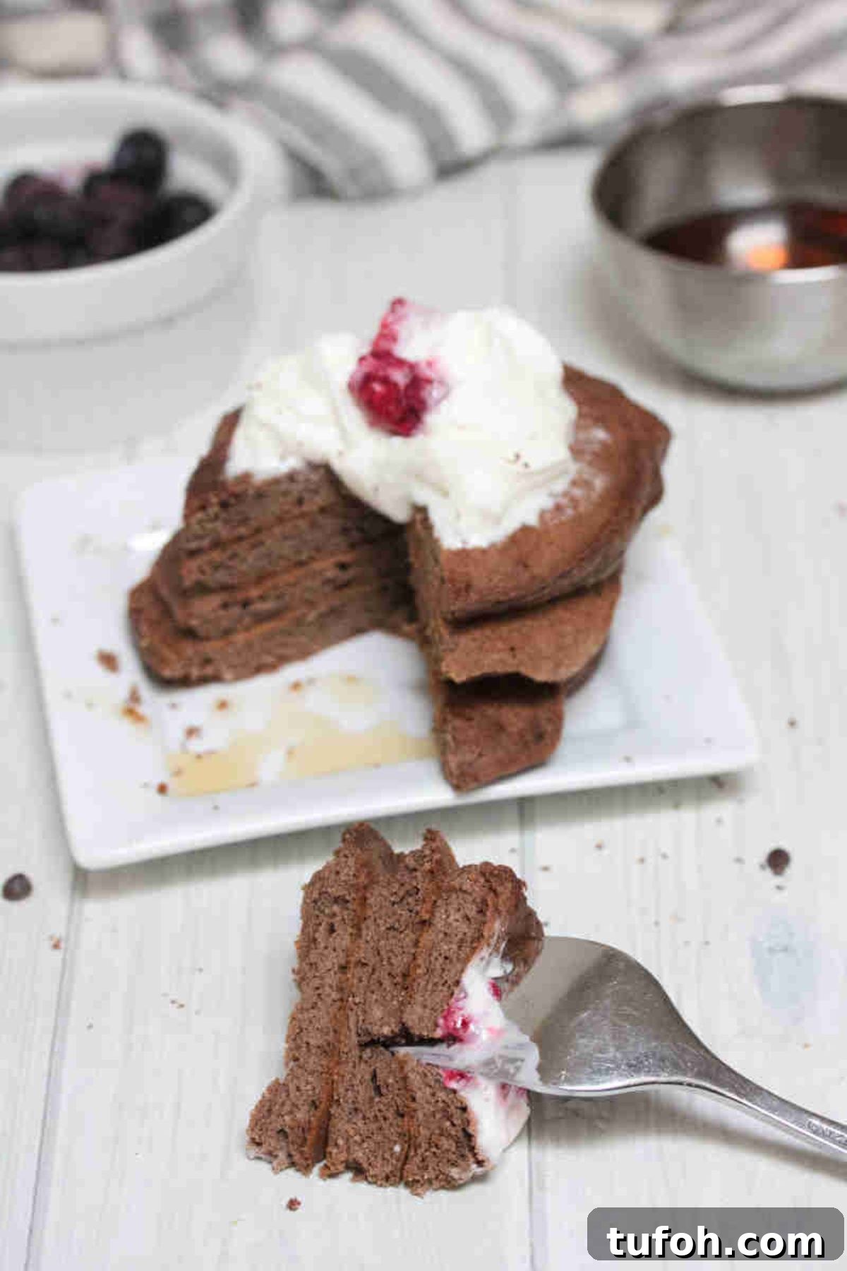 Keto chocolate pancakes stacked on a white plate and whipped cream and raspberries on top, a slice out of the pancakes on the fork in front of the plate.